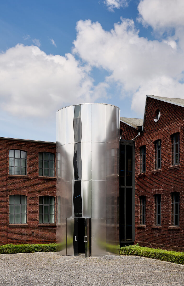 Een bakstenen fabrieksgebouw met een ronde, verspiegelde stalen entreetoren ertussen.