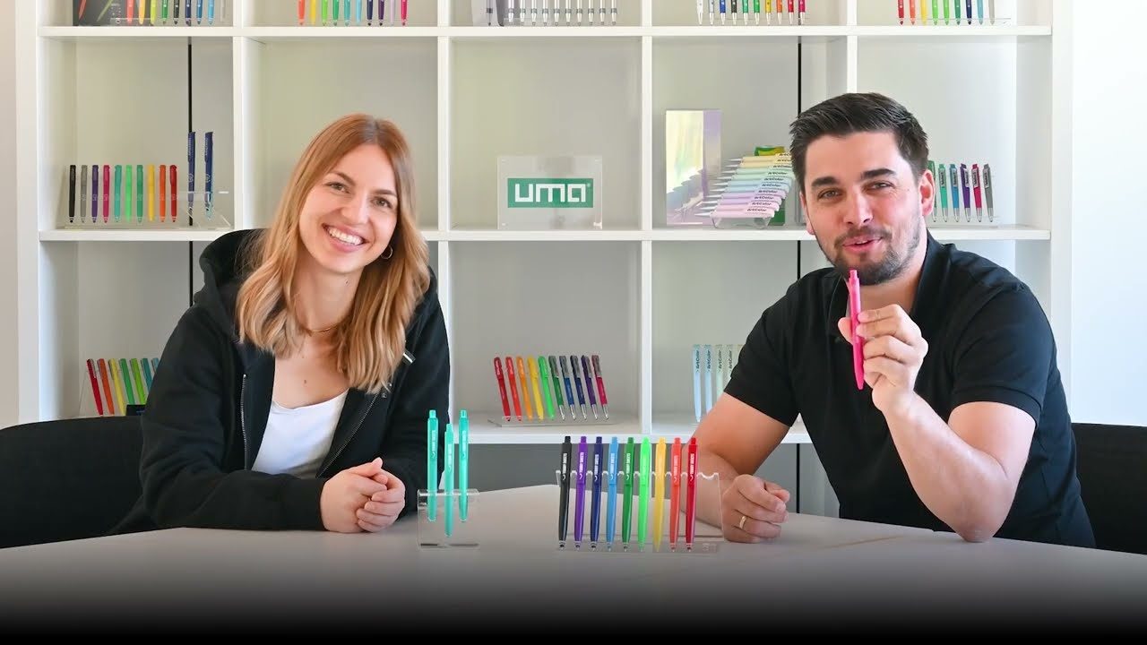Stilstaand beeld uit de video: twee medewerkers zitten lachend aan een tafel met standaards vol gekleurde balpennen voor zich, in een showroom met wandkasten vol pennen en het uma-logo op een van de planken.
